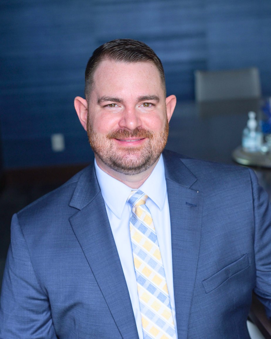 The founder and lead coach of Day One Agent Academy — a professional real estate coach in a blue suit, smiling in a navy office — who built the program to give agents a relationship-based path to success.